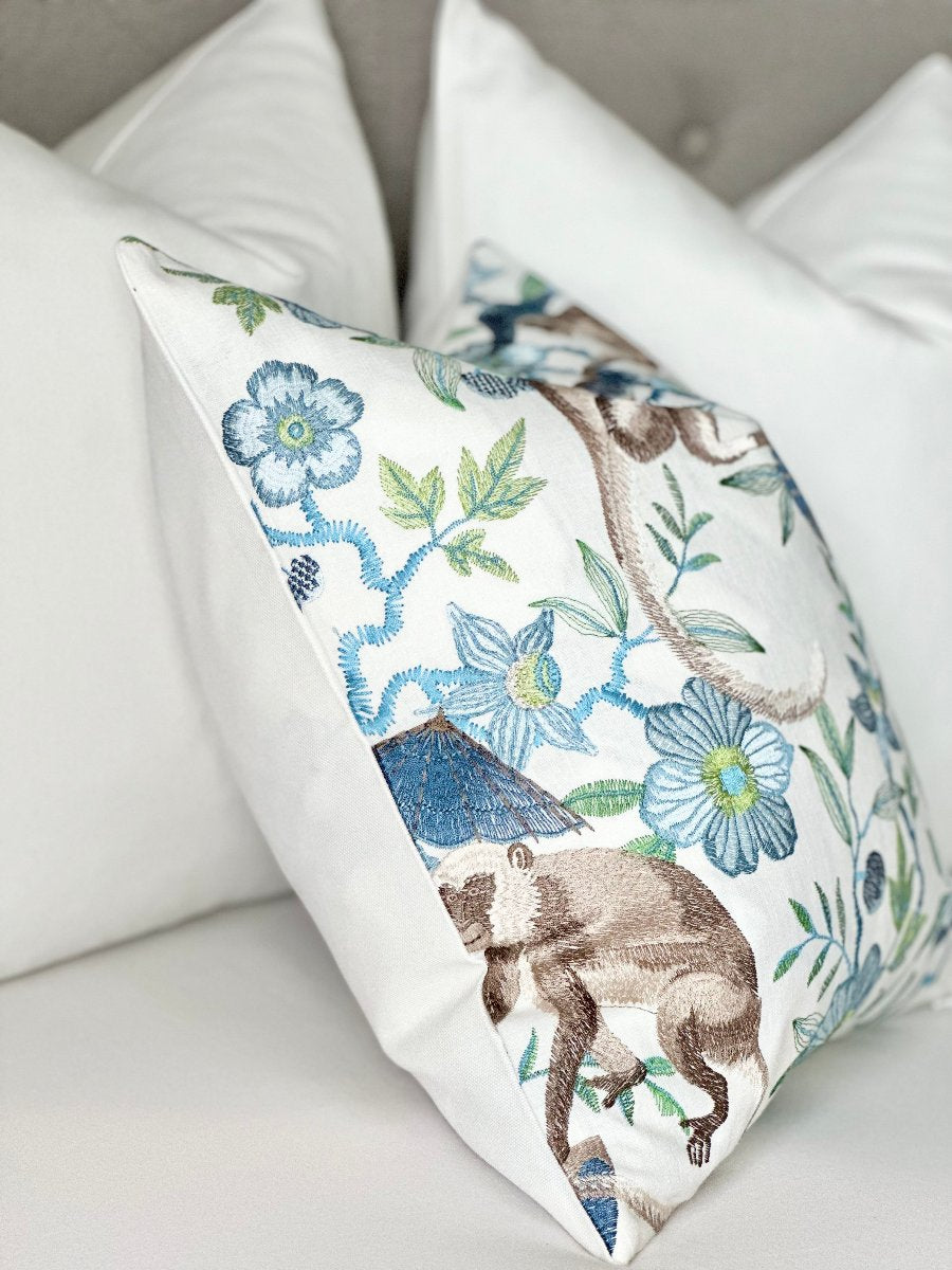 Designer pillow cover featuring cheeky monkeys holding umbrellas with a blue floral embroidery pattern on a saturated ivory backdrop. This unique pillow adds a playful touch to modern, chinoiserie, or blue-themed interiors, serving as a striking accent piece in any space.